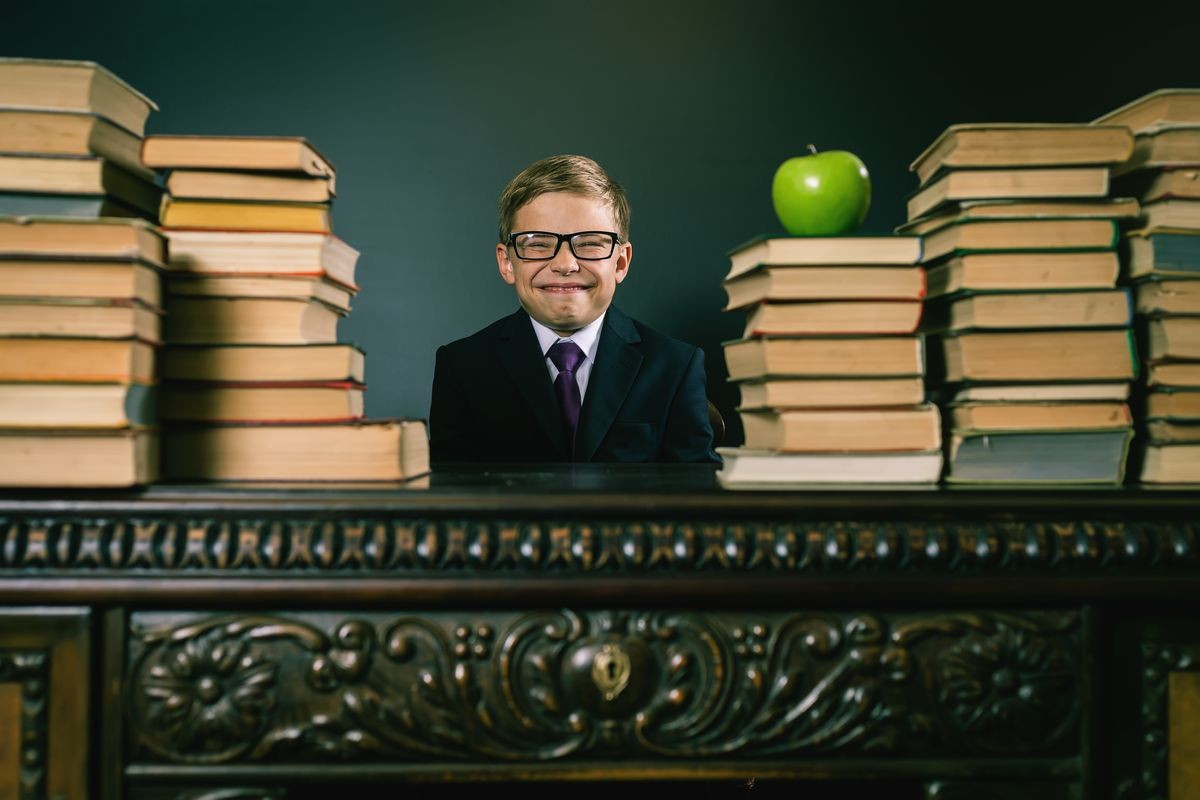 Cunning school boy sitting at the table with many books and one green apple. Smiling child dressed in school uniform and glasses. Blackboard. Student. Concept of education. Looking at camera